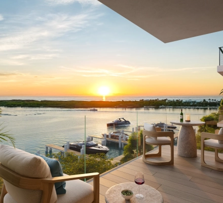 Rendering of a covered wood-decked terrace with a cushioned lounge chair, glass of red wine, bistro table and two chairs behind a glass railing, overlooking docked boats, the Intracoastal Waterway and the sun rising over the ocean.