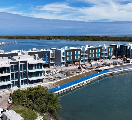 Aerial view of modern luxury waterfront townhomes under construction beside turquoise water and marina.