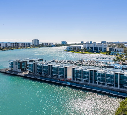 Aerial view of modern luxury waterfront townhomes under construction beside turquoise water and marina.