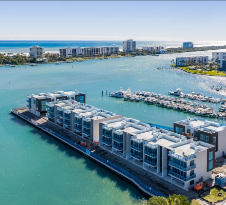 Aerial view of modern luxury waterfront townhomes under construction beside turquoise water and marina.
