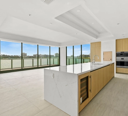 Modern waterfront kitchen with marble island, wood cabinets, and floor-to-ceiling balcony windows.
