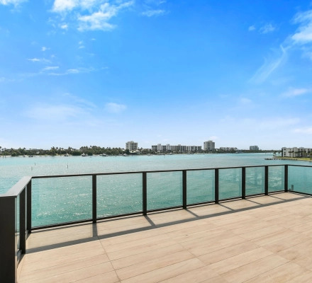 Large tiled terrace with glass railing overlooking turquoise bay water and distant buildings.