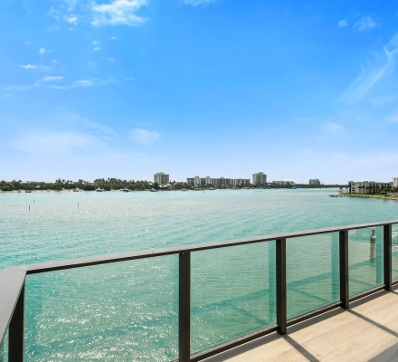 Balcony with glass railing and beige tile overlooking sparkling turquoise water and shoreline.